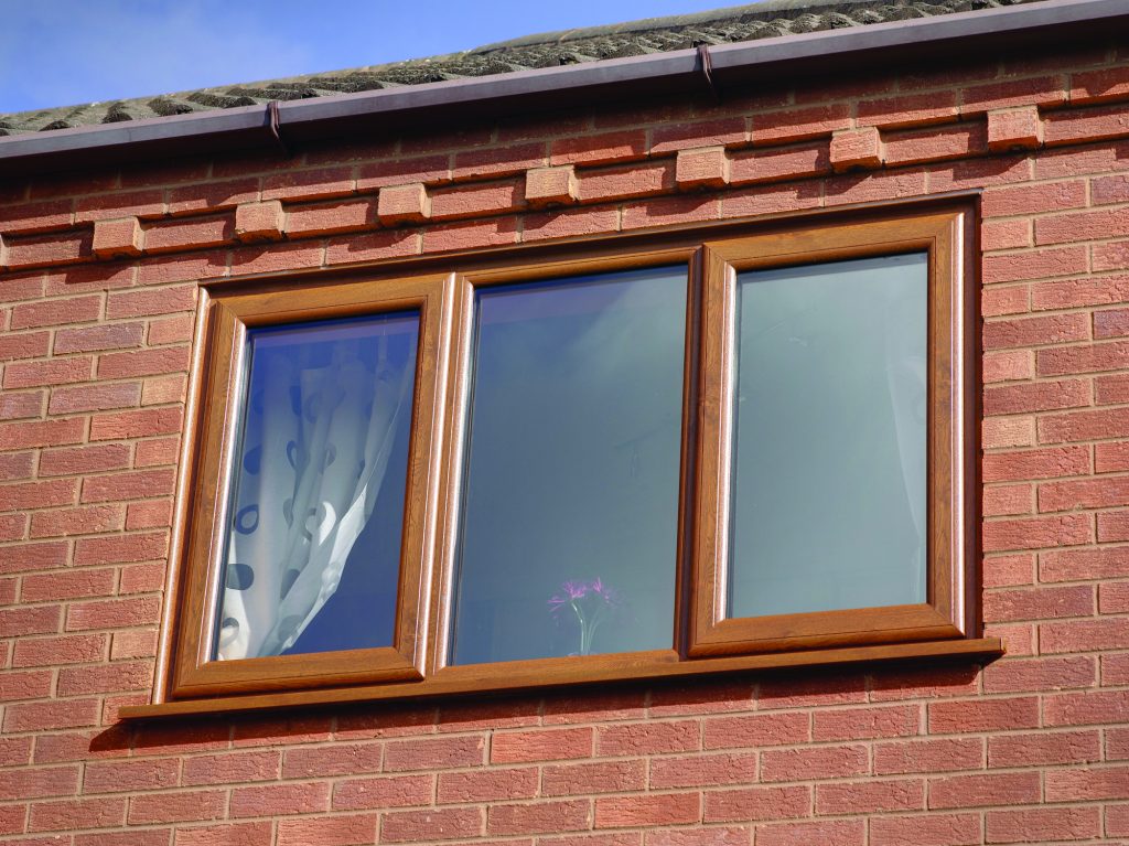 A triple-panel window with a warm oak finish, set in a red brick wall under a slate roof. The leftmost pane features a patterned frosted glass design, while the centre and right panes are clear, with a small, vibrant purple flower visible on the windowsill, adding a splash of colour to the scene.