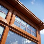 A close-up view of a brown uPVC window with a classic design, featuring multiple panes with white grid inserts. The window is set into a textured, cream-colored exterior wall and is topped with a matching brown gutter. The sky reflected in the glass suggests a clear, sunny day.