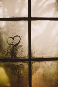 A heart shape is drawn on a misty, condensation-covered window with multiple panes. Sunlight filters softly through the window, casting a warm, diffused light and highlighting the heart, evoking a sense of warmth and affection.