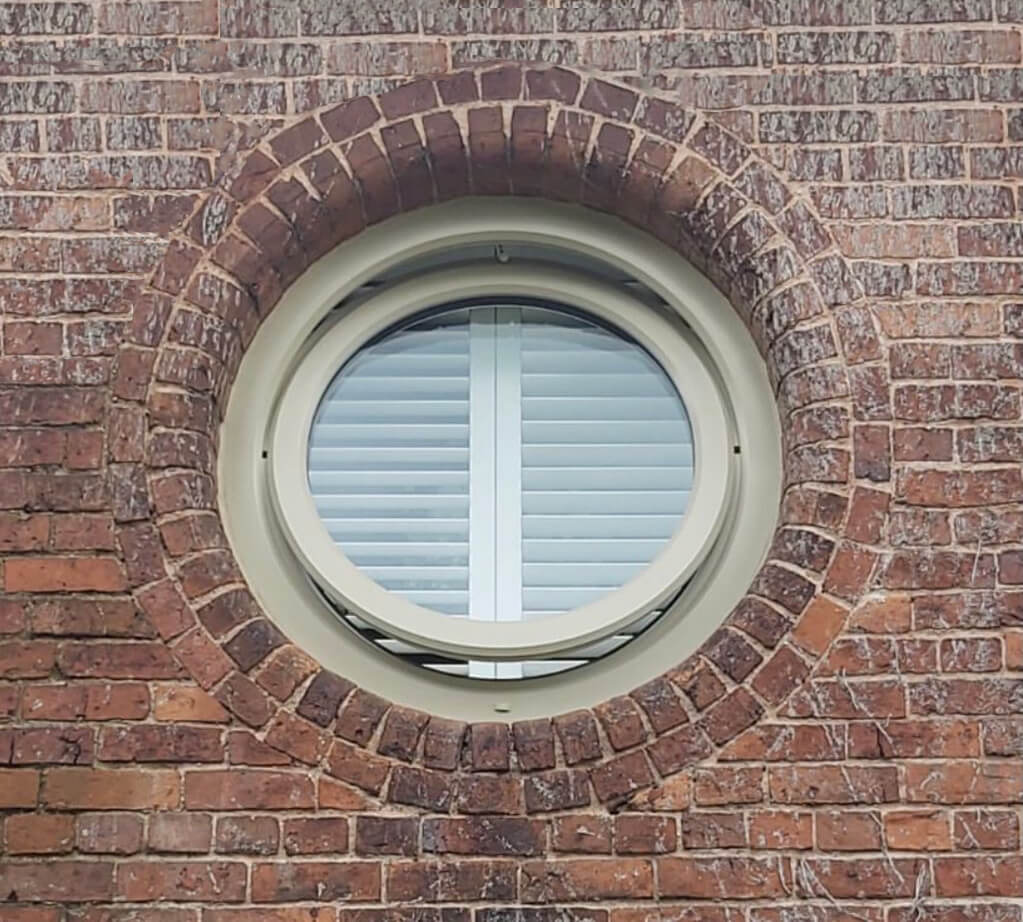 A round cream-framed window set into a red brick wall, featuring internal shutters for privacy and insulation.