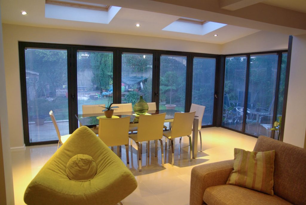 A modern dining space with black aluminium bifold doors fitted with integral blinds, leading to a garden.