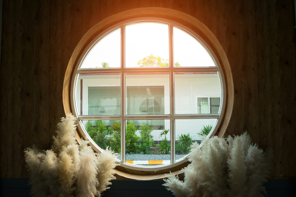 A circular window with decorative grilles, providing a framed view of a modern home with greenery outside.