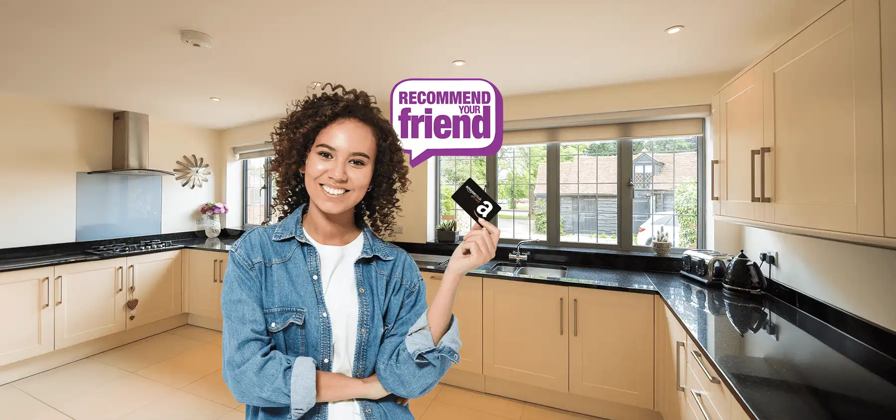Smiling woman holding a Amazon Gift Card standing in a modern kitchen with cream units and black countertops. A purple speech bubble above her head reads 'Refer a friend