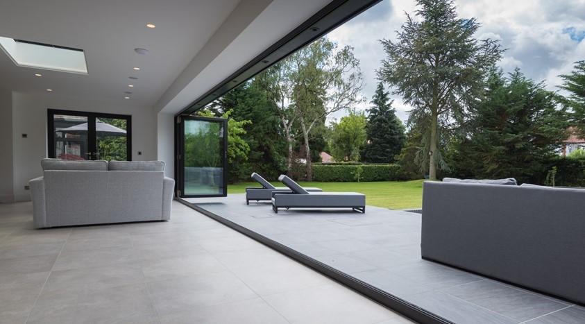 A contemporary living room with an OB-72 bi-folding door configuration (3+0), fully opened to reveal an expansive, manicured garden with lush greenery and tall trees. The room features modern grey sofas and ample natural light from the large glass doors and skylight, creating a seamless connection between indoor and outdoor living spaces.